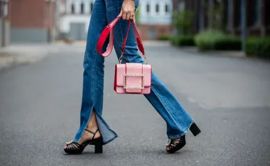 Woman in bootcut jeans and heels holding a pink handbag with a sparkling buckle, walking on a street.