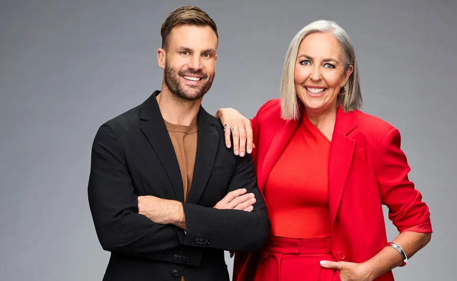 Two people smiling, one in a black suit and the other in a red outfit, against a gray background.