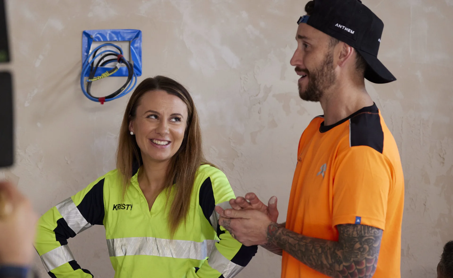 Two people in work attire sharing a light moment; one in a neon jacket with "Kristy" written on it, the other in orange.
