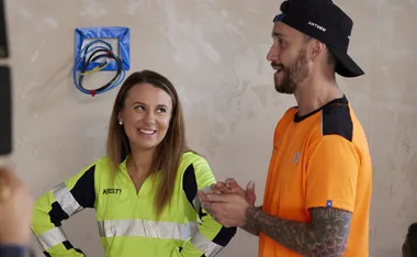 Two people in work attire sharing a light moment; one in a neon jacket with "Kristy" written on it, the other in orange.