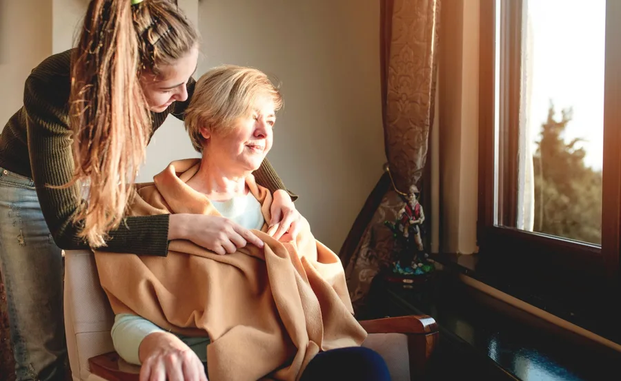 Young woman caring for an older woman, both smiling, near a sunlit window.
