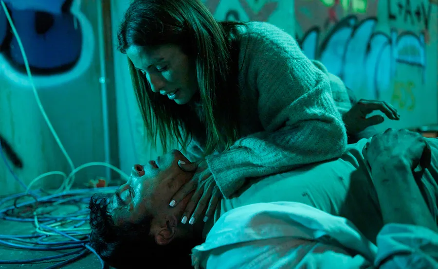 A woman leans over an unconscious man on a graffiti-covered floor, both illuminated by greenish-blue light.