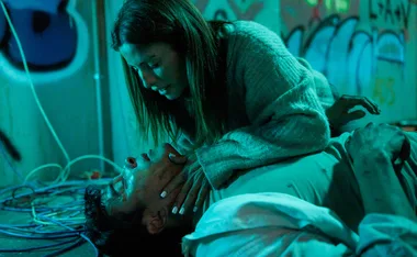 A woman leans over an unconscious man on a graffiti-covered floor, both illuminated by greenish-blue light.