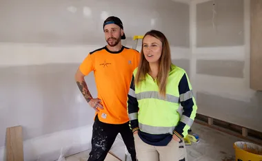 Two people in construction attire stand in a partially built room, appearing to discuss or plan something.