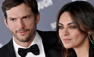 A couple smiling at an event, with the man in a tuxedo and the woman with long hair and earrings.