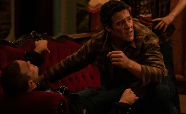 A concerned man kneels over another man lying on a red couch in a dimly lit room.