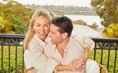 Couple laughing and embracing on a balcony with greenery and water in the background.