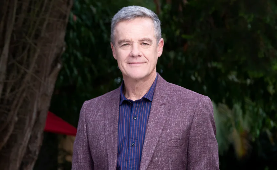 Man with short gray hair wearing a purple blazer stands outdoors with trees in the background.