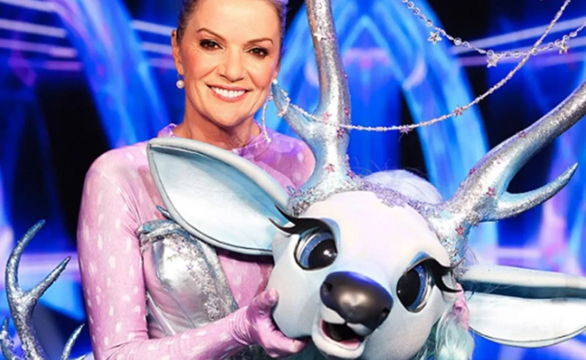 A woman smiles beside the Fawn character head from The Masked Singer Australia 2023, featuring silver antlers and blue background.