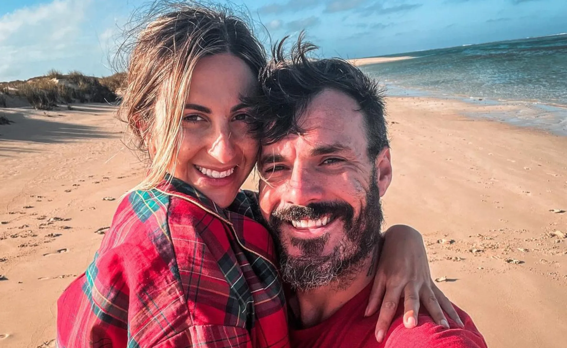 A smiling couple embracing on a sandy beach with the ocean in the background.