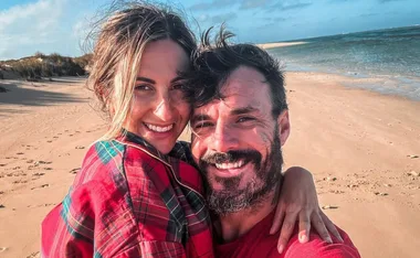 A smiling couple embracing on a sandy beach with the ocean in the background.