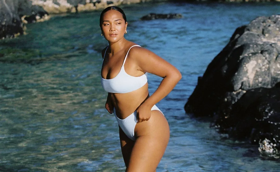 Model in sustainable swimwear stands by ocean rocks under clear sky.