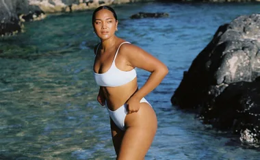 Model in sustainable swimwear stands by ocean rocks under clear sky.