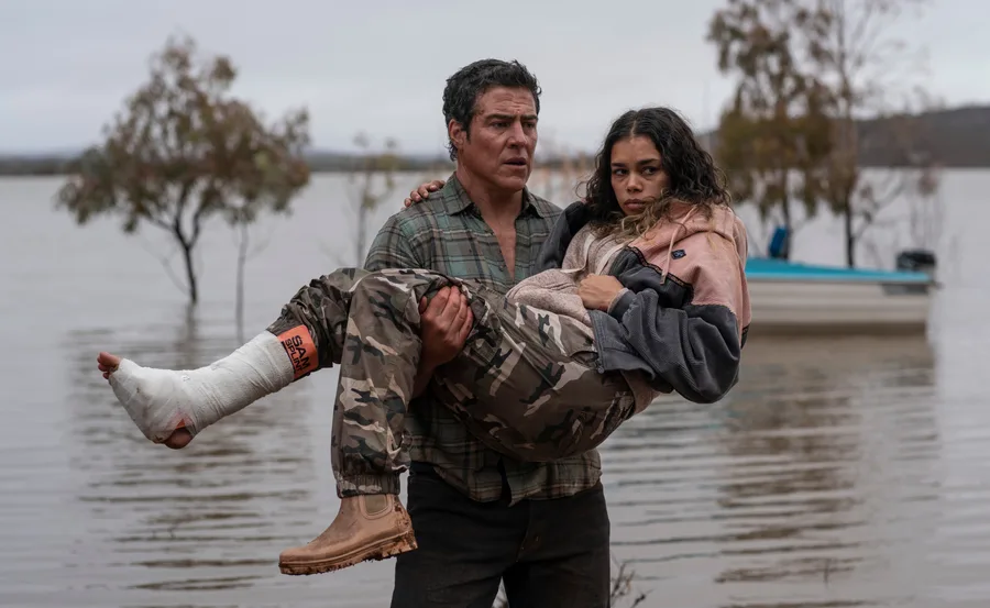 Man carrying woman with injured leg by a flooded area, boat and trees in the background.
