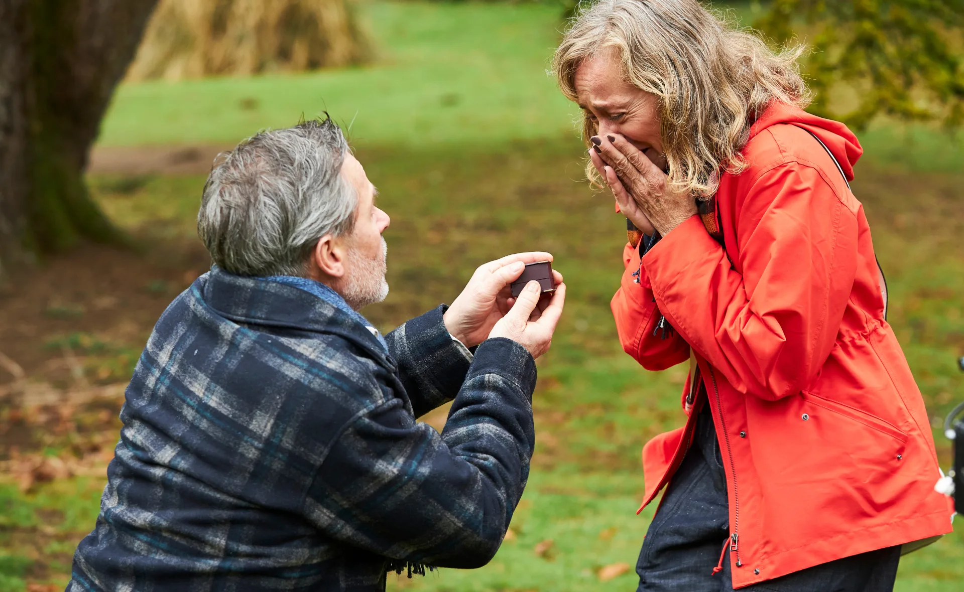 Man kneeling and proposing with a ring to a surprised woman in a red coat outdoors.