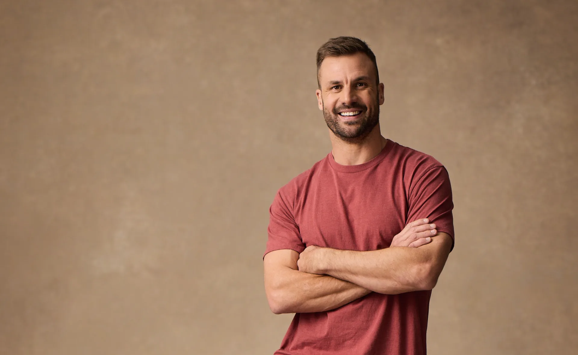 Man in a red shirt standing with arms crossed, smiling against a beige background.