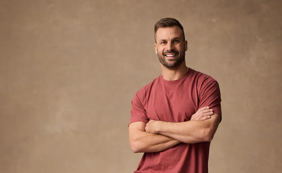 Man in a red shirt standing with arms crossed, smiling against a beige background.