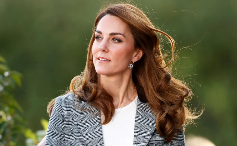 Kate Middleton with flowing brown hair, wearing a plaid jacket, outdoors with a blurred green background.