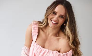 A smiling woman with long, wavy hair in a pink off-shoulder dress against a light background.