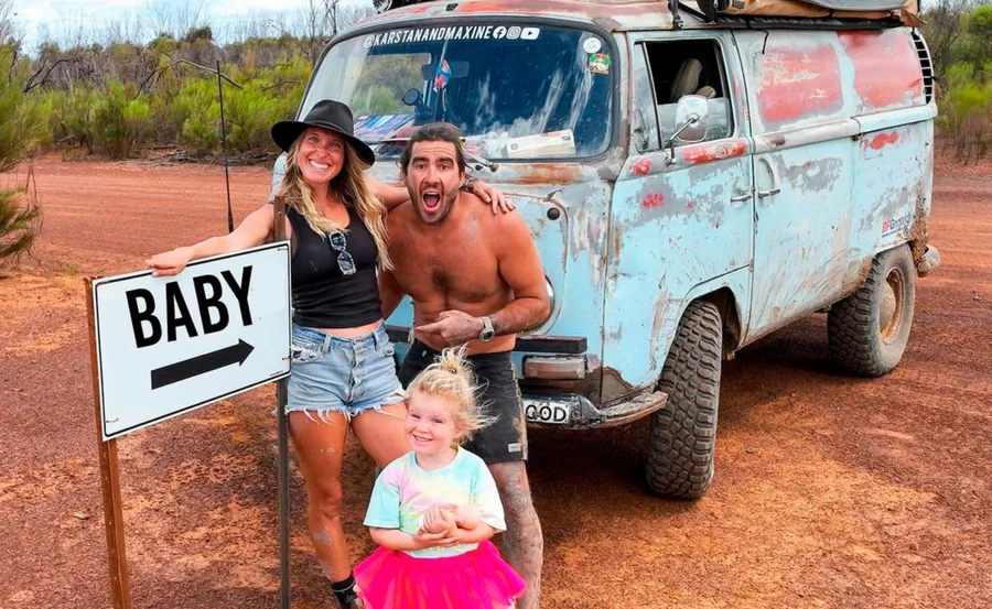 Family posing cheerfully beside a "BABY" sign and a vintage van in a rural setting.