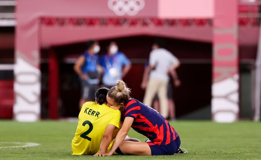 Two soccer players, one in a yellow jersey with "Kerr" on the back, sit on the field, embracing gently.