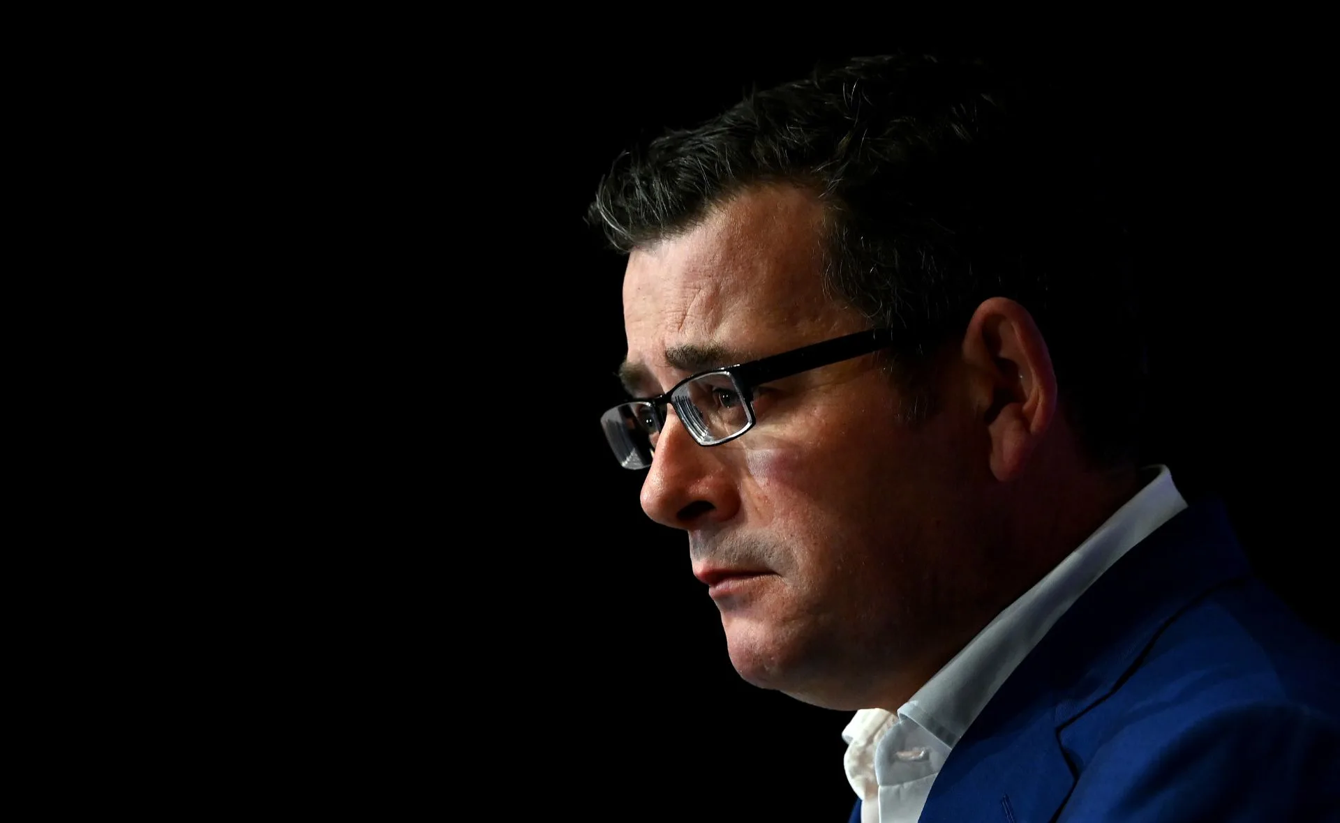 A man in a dark suit and glasses against a black background, appearing thoughtful and serious.