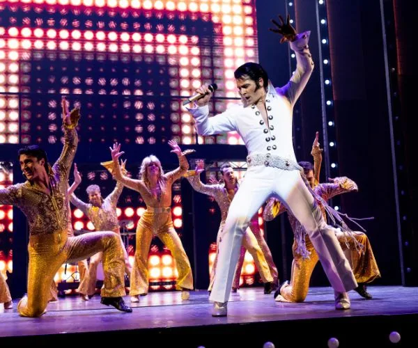 Elvis impersonator performing energetically on stage with dancers in gold outfits and bright lights in the background.
