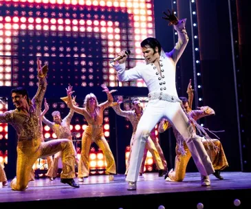 Elvis impersonator performing energetically on stage with dancers in gold outfits and bright lights in the background.