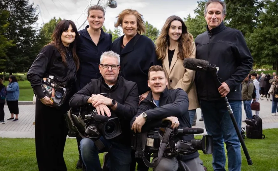 Group photo of seven individuals, including camera crew, outdoors in a park setting with trees and a Ferris wheel.