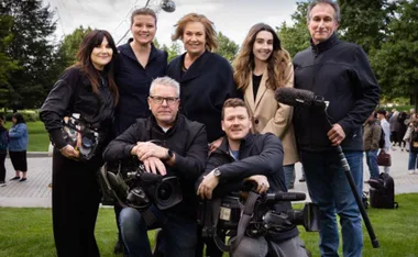 Group photo of seven individuals, including camera crew, outdoors in a park setting with trees and a Ferris wheel.