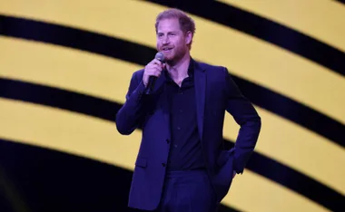 A man in a dark suit speaks into a microphone with a yellow and black striped background.