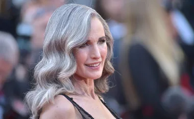 A person with elegant long grey hair smiles, standing in a formal setting with a blurred crowd in the background.