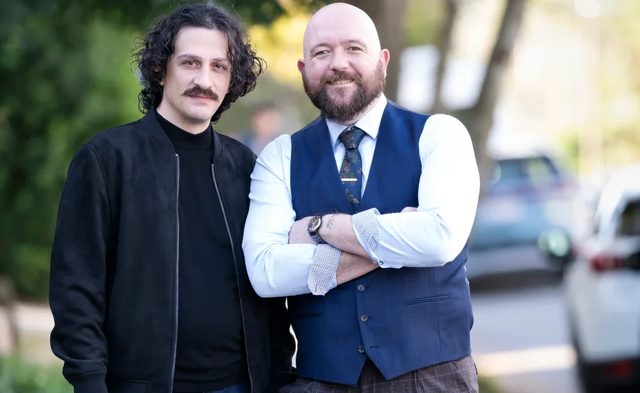Two men posing outside; one in a black jacket, the other in a blue vest, arms crossed, smiling.