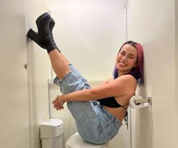 A person with colorful hair sits playfully on a toilet, legs up, wearing denim and high heels, smiling in a small restroom.