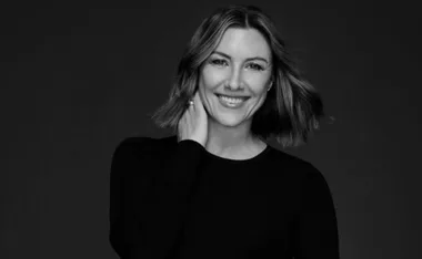A woman with medium-length hair in a black top smiles, holding her neck in a poised, professional portrait.