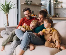 Family relaxing on couch at home, parents with two young children enjoying time together in a cozy living room.