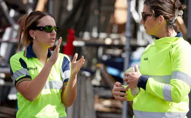 Two women in high-visibility shirts, engaged in conversation on a construction site.