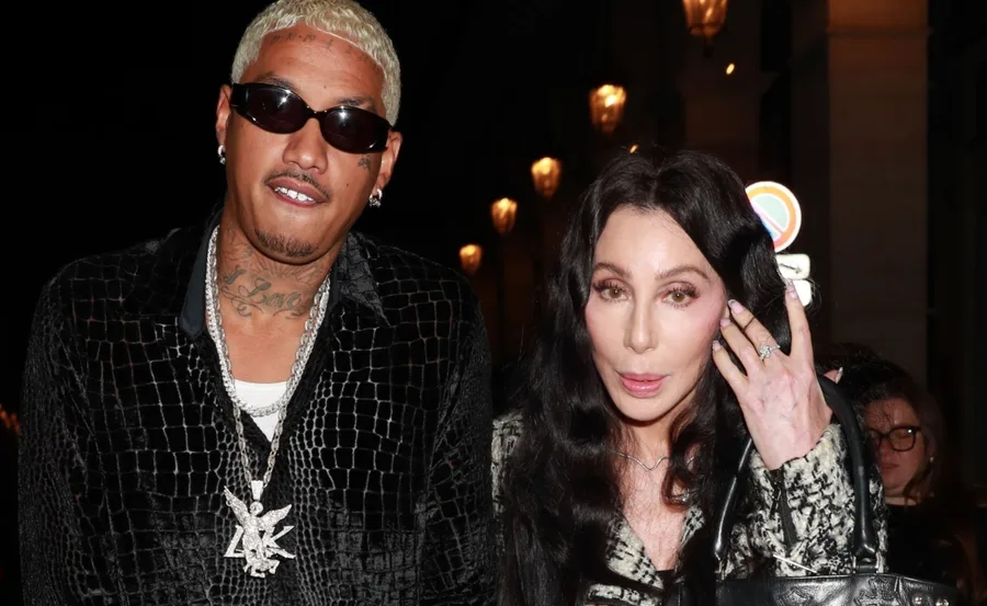 Two individuals posing outdoors at night; one with sunglasses and tattoos, the other with long black hair carrying a bag.
