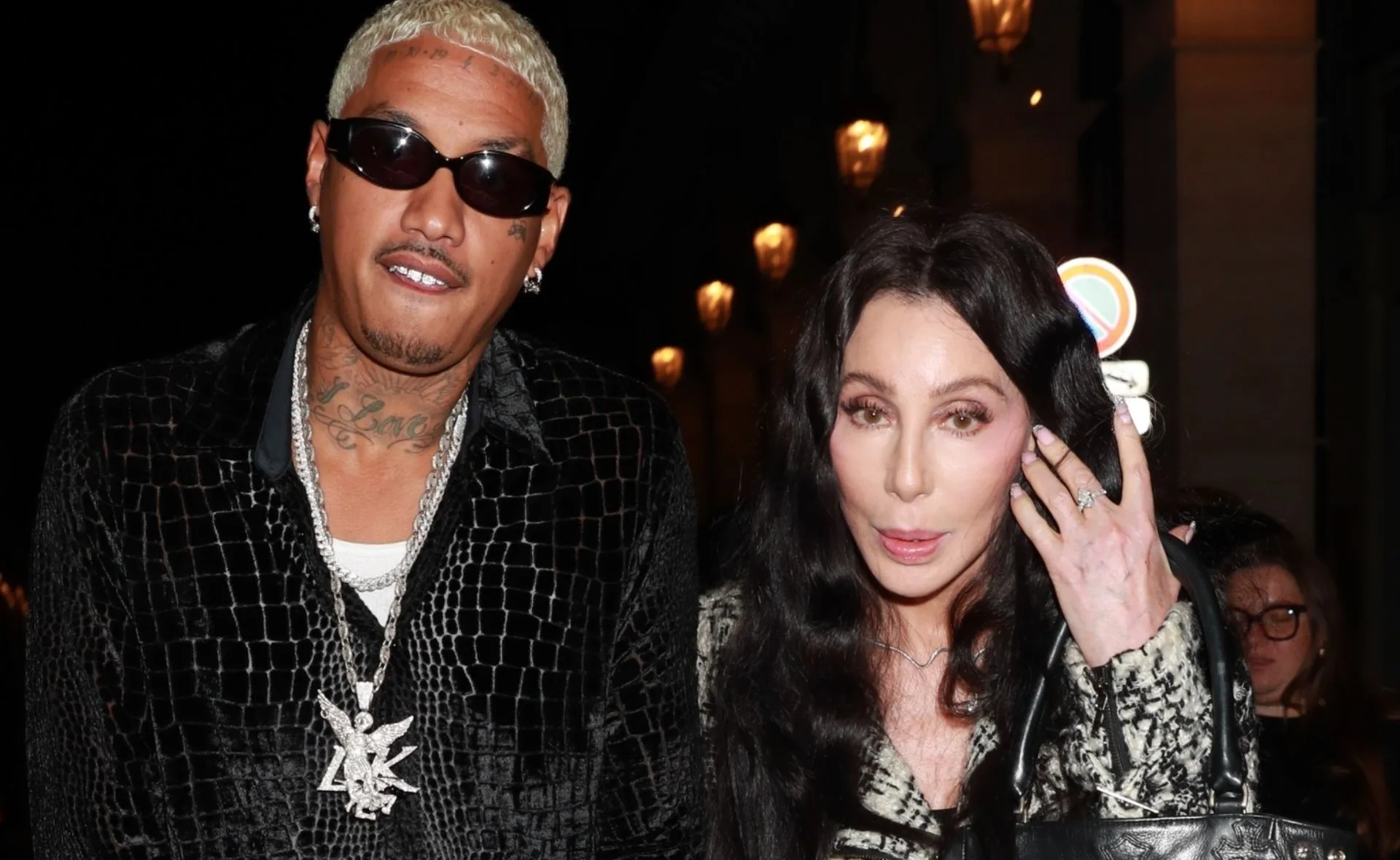 Two individuals posing outdoors at night; one with sunglasses and tattoos, the other with long black hair carrying a bag.