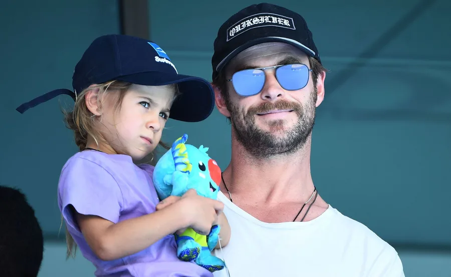 Man in sunglasses holding a child with a baseball cap and a stuffed toy.