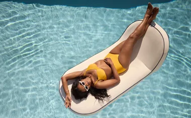 Woman in yellow bikini relaxing on a pool float, wearing sunglasses, surrounded by clear blue water.