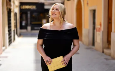 Smiling woman in a black off-shoulder dress holding a yellow clutch, standing on a street.