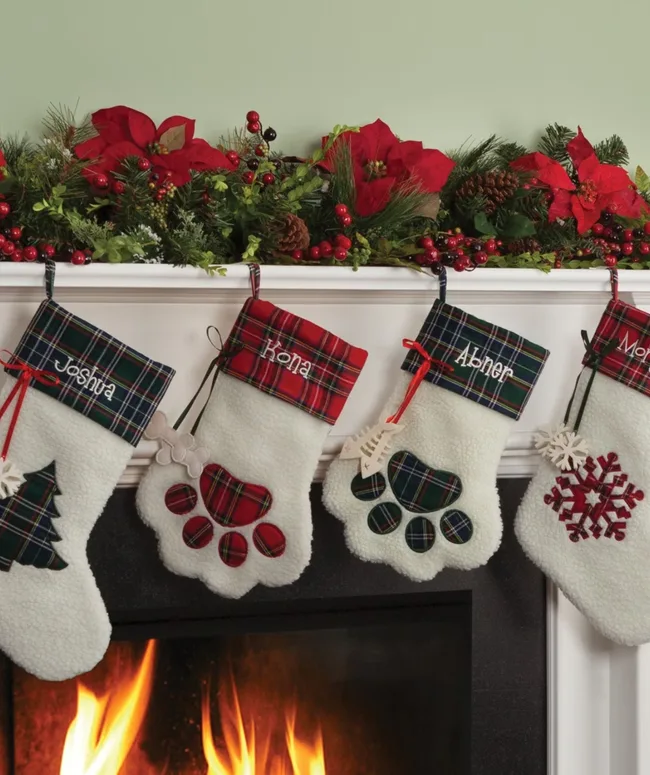 Four paw-shaped Christmas stockings with names, hung on a mantel with garland and poinsettias, above a fire.