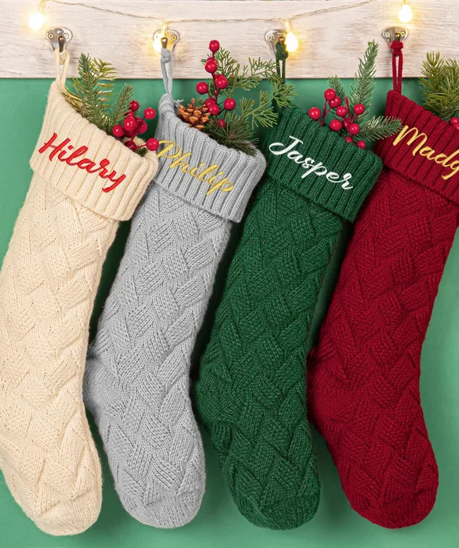 Four knitted Christmas stockings in beige, gray, green, and red with names Hilary, Philip, Jasper, and Madge.