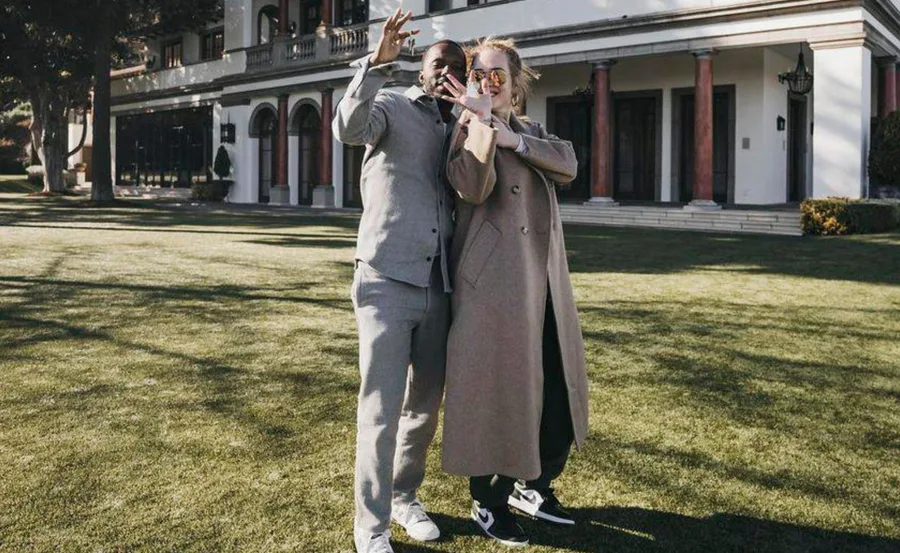 Two people smiling and posing in front of a large house, gesturing with their hands.