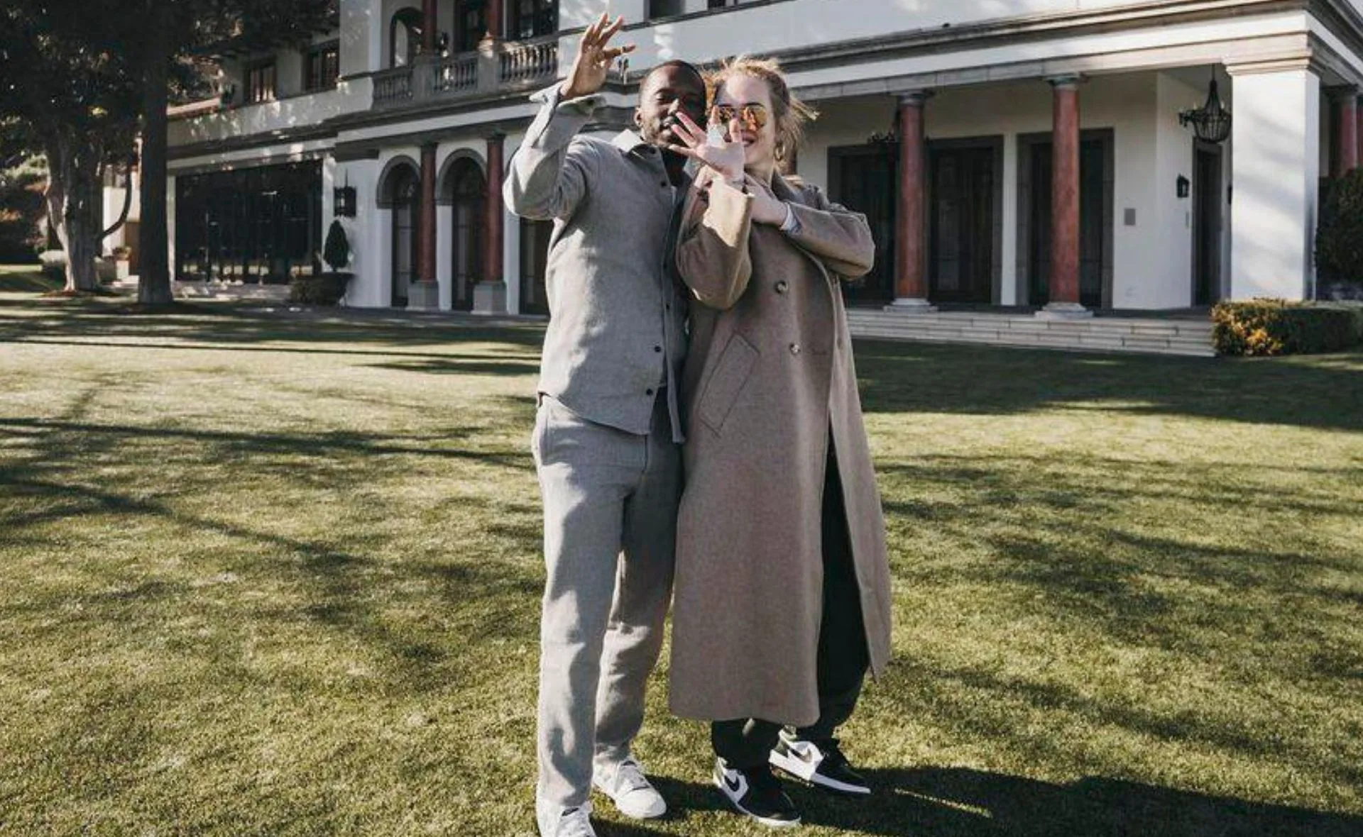 Two people smiling and posing in front of a large house, gesturing with their hands.