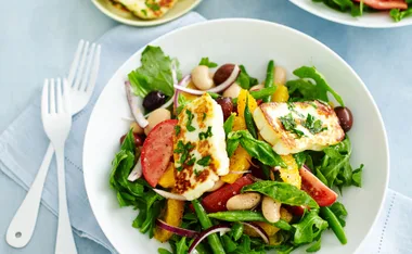 Mediterranean salad with grilled halloumi, arugula, tomatoes, orange slices, beans, and olives in a white bowl.