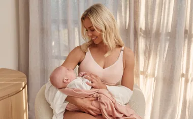 Woman in a pink maternity bra cradles a baby, smiling, in a softly lit room with curtains.