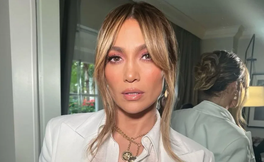 A woman with light brown hair in a white outfit, wearing pink eye makeup, poses indoors in front of a mirror.