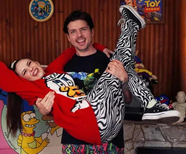 A smiling couple in vibrant outfits; the man holds the woman, surrounded by colorful decor and pop culture posters.